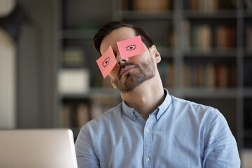 Exhausted young Caucasian man worker have sticker pads on eyes sleeping near computer in office. Tired millennial male employee fall asleep doze off at workplace, feel overwhelmed drained at work.