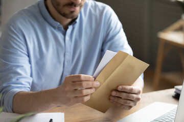 Crop close up of man sit at desk open envelope with paper letter or correspondence at office. Male worker get postal paperwork or notice notification at workplace, receive message or invitation. - obrazy, fototapety, plakaty