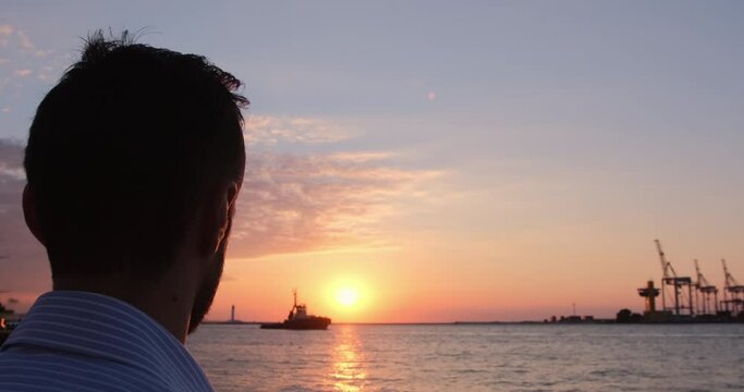 A man watches as a small ship sails past the sun in the early morning at the seaport.