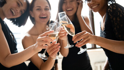Close up focus on glasses with champagne or white wine joined together, happy beautiful multiracial female friends in black cocktail dresses celebrating birthday, feeling excited together at party.