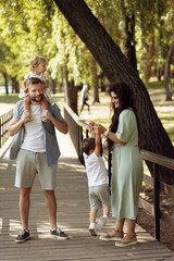 Fototapeta premium Happy parents with little kids at the park, cute daughter sitting on fathers shoulders, lovely mother lift in air joyful son, enjoy happy weekend outdoors, parenting concept