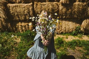 Young girl in rubber boots with flowers standing against the background of straw bales on country...