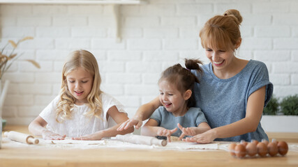 Having fun in the kitchen. Happy mommy and daughters flattening dough on wide wooden counter in cozy modern kitchen. Cheerful young mother teaching her curious 5 and 8 year olds to make pastry dough