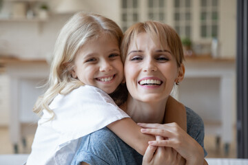 Happy preschool daughter embracing her good looking young mommy, both looking at camera and...