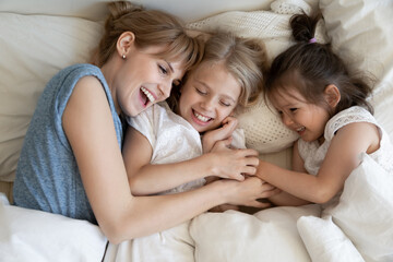 Cheerful mommy in her 30s cuddling with adorable daughters lying in comfortable bed under soft white duvet. Mother and 5 and 8 year old children enjoying weekend at home and lazing on soft pillows