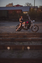 Beautiful sexy woman in lace bodysuit and high heels sits on a motorcycle on an industrial background