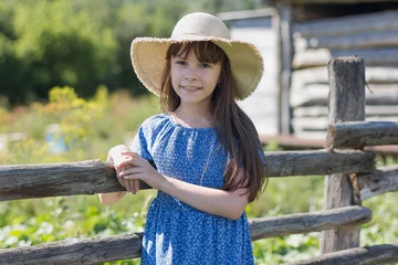 Fotobehang Een jong meisje met lang haar en in een hoed en een jurk staat in de buurt van houten fantasie. Portret van schattig kind op een dorp. Meisje poseren glimlachen en kijken naar de camera. © Albert Ziganshin