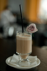 A cup of hot latte coffee in transparent glass on white plate decorated with  a small purple rose.