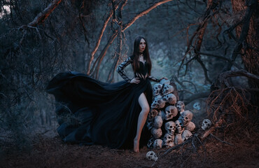 A gothic beautiful young woman in a black dress stands near a mountain of human skulls. Fashion...