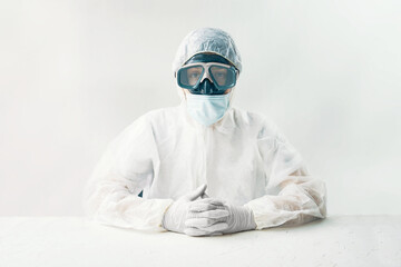 Man in white hazmat on white background studio. Person in a protective suit mask and gloves sits at a table and looks at the camera. 
