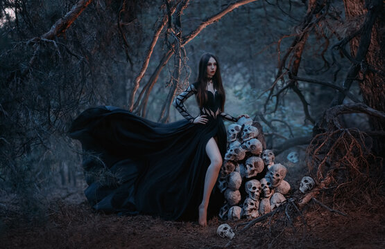 A gothic beautiful young woman in a black dress stands near a mountain of human skulls. Fashion model in the image of the goddess of death in a dark forest of trees. Silk fabric flies in the wind