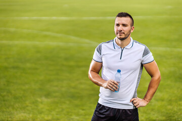 Attractive soccer player in shape standing on the field and holding bottle of water.