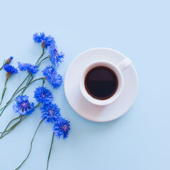 Top view of coffee / hot chocolate cup and a bouquet of blue cornflowers on a light blue background. Summer flat lay. Lifestyle, copy space, mockup, square photo.