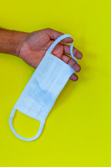 Left hand holding a white face mask on a yellow background. Public health