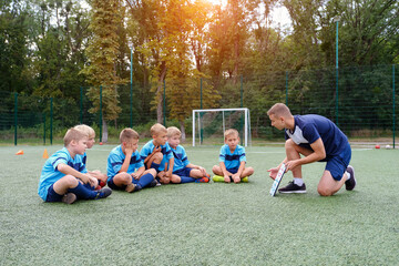 Young coach with clipboard teaches children the strategy of playing on the football field outdoors. theoretical studies in sports training
