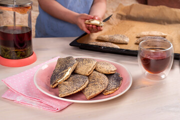 Child hands holding traditional Russian sweet shortcrust pastry with quark or cottage cheese and raisins, sochni or sochniki pies or pirozhki on pink plate with tea in the morning on wooden table