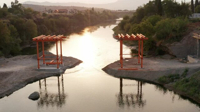 Bridge construction: metal scaffolding used as temporary support for the deck of a small pedestrian bridge. Pair of iron frames on the banks of a narrow river at sunset. 