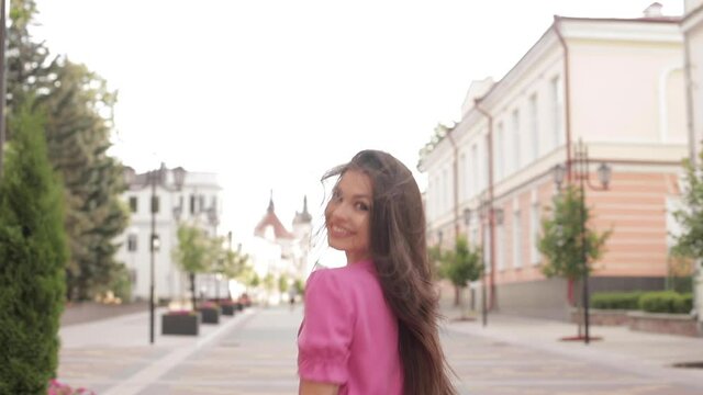 Smiling beautiful young woman with loose long dark hair holding a flower in her hand while running in the city. Tracking shot