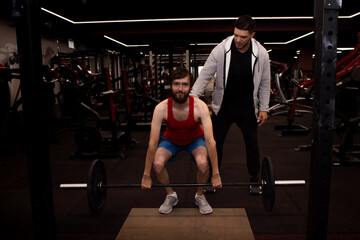 Fototapeta na wymiar Young man doing exercises with a personal trainer as he lifts a barbell weight to chest height in gym