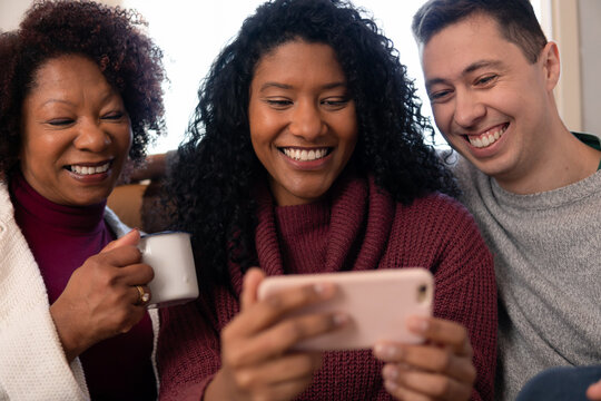 Cheerful brazilian black woman with mother and boyfriend sharing video on a smart phone Inside the house in living room. Relationship, leisure, enjoyment, aging concept..