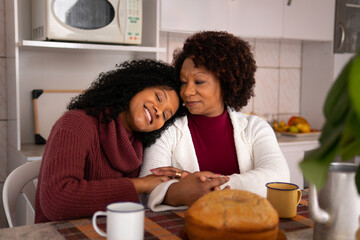 Lovely latin black woman with mother embracing each other at kitchen table, inside. Affectionate, bonding, love, generation concept..
