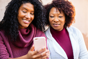 Cheerful latin adult daughter and mother with smart cell phone looking at cell phone screen outside...