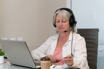 Serious older female doctor with headset taking an online medical appointment in doctors office...