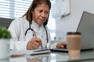 Fototapeta na wymiar Focused old aged female doctor with pen taking notes and looking at laptop screen at medical clinic health, medicalcare, care concept..