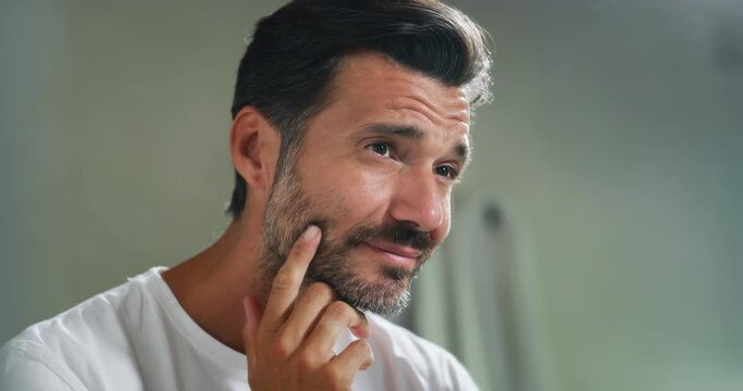 An young smiling man is applying after cleaning his face day or night cream to take care of skin behind a mirror in a bathroom.