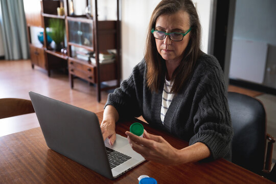 Woman wearing glasses using laptop while holding empty medication container at home