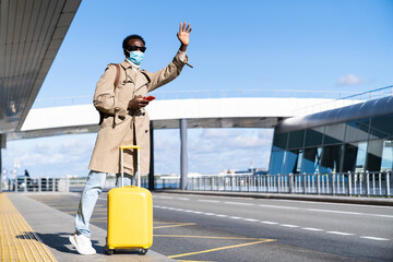 Afro-American millennial man with yellow suitcase stands in airport terminal, using phone, calling taxi, raising hand, wear face medical mask to protect yourself from contact with flu virus, covid-19