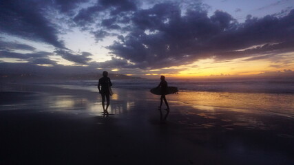 Silueta de dos amigos caminando con tabla de surf en la orilla de la playa con la última luz del atardecer en el horizonte en un día con sol, nubes y montañas con pueblo al fondo