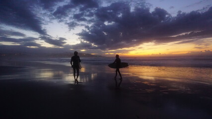 Fototapeta premium Silueta de dos amigos caminando con tabla de surf en la orilla de la playa con la última luz del atardecer en el horizonte en un día con sol, nubes y montañas con pueblo al fondo