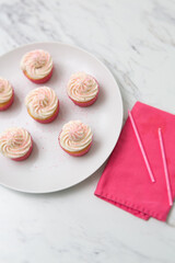 Top View of Vanilla Cupcakes with Vanilla Icing in Pink Paper Cups with Pink Sprinkles on a White Plate on White Countertop with Pink Napkin Beside