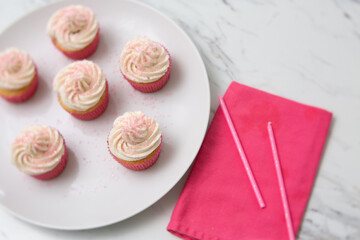 Top View of Vanilla Cupcakes with Vanilla Icing in Pink Paper Cups with Pink Sprinkles on a White Plate on White Countertop with Pink Napkin Beside