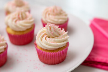 Close Up of Vanilla Cupcakes with Vanilla Icing in Pink Paper Cups with Pink Sprinkles on a White Plate on White Countertop with Pink Napkin Beside