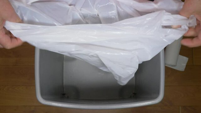 Closeup POV overhead shot of a man&rsquo;s hands putting a clean plastic bag from a roll, for kitchen food waste, over the rim of a small bin / trash can, then using a hand to open it up, ready for use.