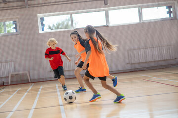 Three kids in sportswear playing indoors football in the gym and looking excited © zinkevych