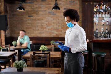 Black waitress with protective face mask and gloves working on touchpad in a cafe.