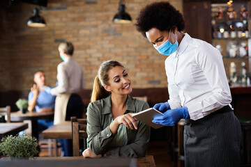 Black waitress with face mask taking order on touchpad from a customer in a cafe.