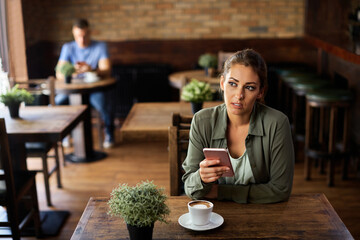 Pensive woman texting on mobile phone while drinking coffee in a cafe.