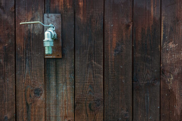A silver-colored metal water tap is placed on a brown wooden wall. The boards are discolored and cracked with age. Some of them are wet. Background. Texture.