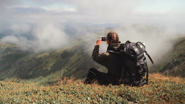 young beautiful woman sits on the top of the mountain and takes a beautiful photo on her phone