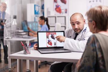 Fototapeta na wymiar Doctor looking at senior woman while pointing at heart diagram in hospital office during consultation. Nurse in blue uniform holding x-ray image.