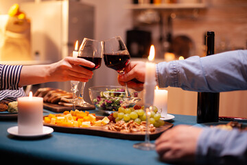 Couple holding glasses with red wine during romantic relationshiop celebration. Happy cheerful young couple dining together in the cozy kitchen, enjoying the meal, celebrating anniversary romantic