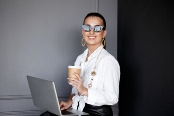 Cheerful girl in white shirt and black pants, sunglasses, siting and working on laptop and holding a cup coffee, over black and grey background.