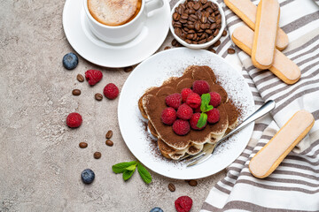 portion of Classic tiramisu dessert with raspberries and cup of coffee on grey concrete background