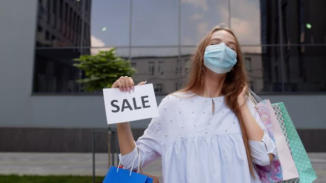 Girl in protective mask with shopping bags showing Sale word inscription during coronavirus pandemic