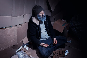 A homeless bearded man sits on boxes on the street and asks for help. Need a homeless person asks for money for food and overnight.
