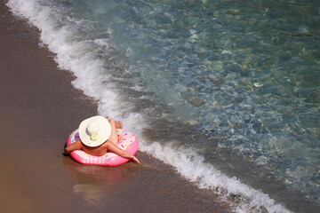 Woman in hat laying on inflatable donut ring on a sand in sea waves, aerial view. Beach vacation, relax and leisure in clean water